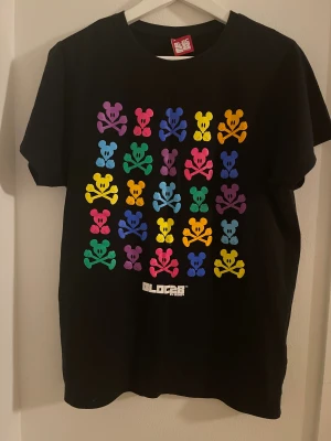 Svart Disney Bloc28 t-shirt med färggrafik - Cool svart t-shirt från Disney Bloc28 med färgglada Mickey Mouse-skallar i regnbågsmönster över hela framsidan. Trycket har starka nyanser av lila, rosa, blå, grön, gul och orange. Klassisk rund hals och korta ärmar. Perfekt för dig som gillar streetstyle och grafiska prints.