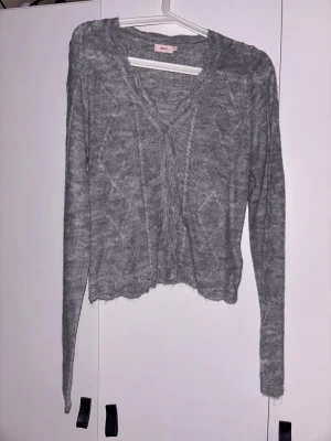 Grå stickad kofta från Nelly - Säljer en grå stickad kofta från Nelly med v-ringad hals och knappar framtill. Koftan har ett diskret mönster och långa ärmar. Den är mjuk och har en lite ruffad kant nedtill för en chill vibe. Perfekt att slänga på över en t-shirt.