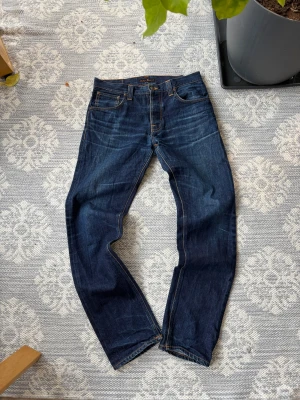 Mörkblå jeans från Nudie Jeans, W33/L34 - Säljer ett par mörkblå jeans från Nudie Jeans med klassisk femficksdesign och kontrastsömmar. Jeansen har rak passform och är tillverkade i slitstark denim av bomull. Snygg detalj med Nudie-loggan på bakfickan och läderpatch i midjan. Lite ljusare färg i midjan men inget hål eller liknande 