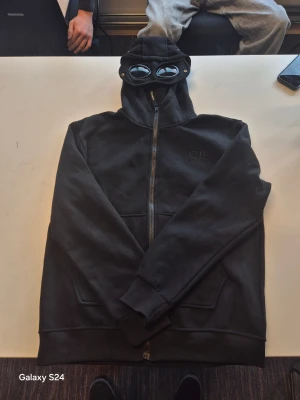 Svart gogglejacka från C.P. Company - Svart gogglejacka från C.P. Company i medium med ikoniska glasögon i huvan. Jackan har dragkedja framtill, två stora fickor och en clean look. Perfekt för dig som vill sticka ut med streetwear-vibe och coola detaljer.