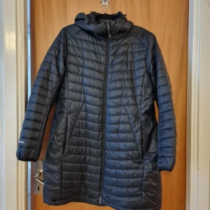 Svart quiltad dunjacka från 67°N Áhkká - Svart lång dunjacka med huva från 67°N Áhkká. Jackan har quiltad design, dragkedja framtill och praktiska fickor med dragkedja. Perfekt för kalla dagar och har en stilren look som funkar till det mesta. Snyggt broderad logga på ärmen.