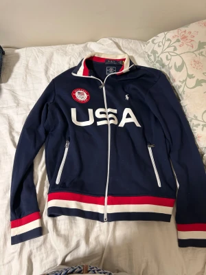 Polo Ralph Lauren tröja  - Snygg marinblå track jacket från Polo Ralph Lauren, officiell USA Olympic Team 2022. Jackan har vita och röda detaljer, dragkedja framtill och på fickorna, samt broderad logga och USA-tryck på bröstet. Ribbad krage och muddar med röd och vit rand.  OPS ⚠️för stor för mig kanske ser konstigt ut 