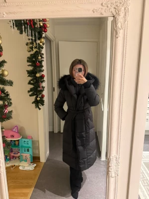 Svart lång dunjacka med riktig päls - Säljer en svart, lång dunjacka med fluffig riktig päls på huvan och bälte i midjan. Jackan har quiltad design, dragkedja och långa ärmar. Perfekt för kalla dagar och ger en snygg siluett med midjebälte. Jackan är från Femenina of sweden. Casablanca modellen stl S