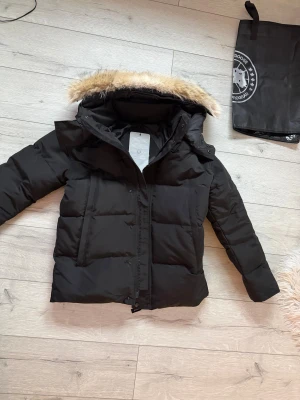 Svart dunjacka från Canada Goose - Svart dunjacka från Canada Goose med pälskantad huva och klassisk logga på ärmen. Jackan är quiltad och har dragkedja framtill. Perfekt för kalla vinterdagar och har en stilren, tidlös look.