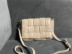 Beige axelväska från Nocila - Snygg beige axelväska från Nocila med flätad design och justerbar axelrem. Väskan har magnetlås, guldfärgade detaljer och ett innerfack med dragkedja. Perfekt storlek för det viktigaste och stilren look som lyfter din outfit.