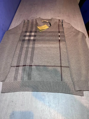 Beige rutig stickad tröja Burberry - Snygg beige stickad tröja från Burberry med klassiskt rutigt mönster i grått och vinrött på framsidan. Tröjan har rund hals, ribbade muddar och lång ärm. Perfekt för dig som gillar stilrena designer och vill ha något som sticker ut lite extra.