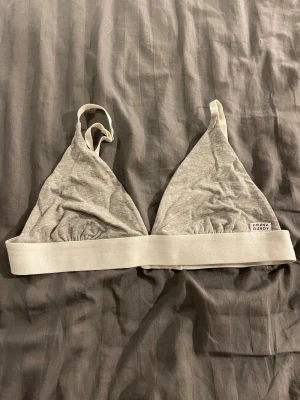 Grå triangeltopp från Frank Dandy - Triangelformad grå topp från Frank Dandy med vita justerbara axelband och bred resår under bysten. Mjuk bomullskänsla och enkel design med liten logotyp på ena sidan. Perfekt för en chill och avslappnad stil.