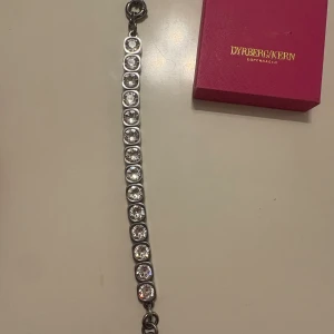 Dyrberg/Kern armband med kristaller - Stilrent armband från Dyrberg/Kern i silver- och guldfärgad metall med stora, gnistrande klara kristaller infattade i fyrkantiga länkar. Armbandet har en chunky kedja och rund bricka med logga. Perfekt statement piece för dig som gillar bling.
