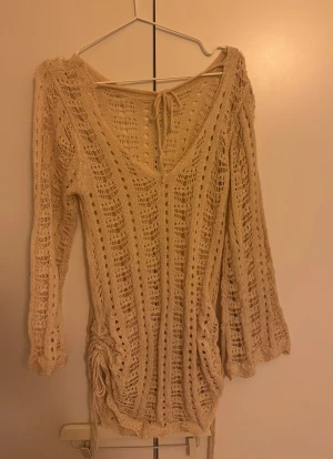 Beige stickad kortklänning med snörning - Beige stickad kortklänning med v-ringning och långa ärmar. Klänningen har ett öppet, virkat mönster och snörningsdetalj i ryggen samt i sidorna nertill. Perfekt för sommarens varma dagar och ger en chill boho-vibe.