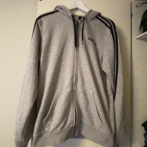 Grå Adidas hoodie med dragkedja - Grå hoodie från Adidas med svarta ränder längs axlarna och ärmarna. Tröjan har huva med snörning, två fickor framtill och hel dragkedja. Perfekt för en sportig och avslappnad stil.