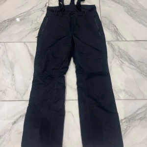 Svarta täckbyxor med hängslen - Svarta täckbyxor med justerbara hängslen och dragkedjeförsedda fickor. Byxorna har knäppning i midjan och förstärkta benslut, perfekta för kalla dagar i backen eller snölek. Snygg och funktionell design med praktiska detaljer.