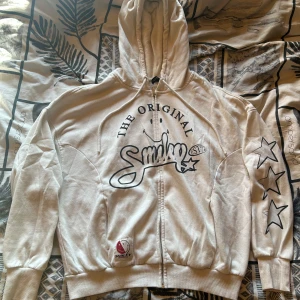 Vit hoodie från Smiley med tryck - Vit hoodie från Smiley med svart tryck framtill och stjärnor på ena ärmen. Tröjan har huva, dragkedja och ribbade muddar. Perfekt för dig som gillar streetstyle och vill sticka ut med coola detaljer.