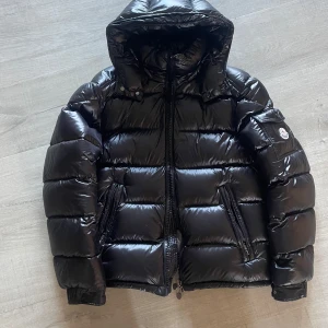 Moncler Maya svart - Säljer en svart, glansig dunjacka från Moncler med avtagbar huva och dragkedja framtill. Jackan har två fickor med dragkedja och Moncler-logga på ärmen. Perfekt för kalla dagar och riktigt snygg streetstil.
