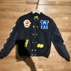 Svart bomberjacka Off-White Kids - Svart bomberjacka från Off-White Kids med broderade patches i blått, gult och orange framtill, samt vit logga på ryggen. Jackan har vita pilar på ärmarna, tryckknappar och ribbade muddar. Materialet är polyester och jackan har en loose passform. Aldrig använt, fick den som present då den var för liten