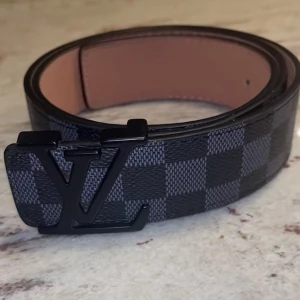 Louis Vuitton rutig svart bälte - Snyggt bälte från Louis Vuitton med klassiskt rutigt mönster i svart och grått. Bältet har en stor LV-logga i svart metall som spänne och är gjort i exklusivt skinn. Perfekt accessoar för att lyfta din outfit.