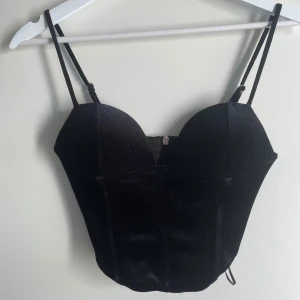 Svart sammetskorsett med smala axelband - Säljer en svart korsettopp i sammet med smala, justerbara axelband och markerade kupor. Toppen har dragkedja bak och en figurnära passform som ger en snygg siluett. Perfekt för att lyfta vilken outfit som helst.