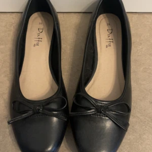 Svarta ballerinaskor från Duffy - Stilrena svarta ballerinaskor från Duffy med rund tå och dekorativ rosett framtill. Skorna har en klassisk design och är tillverkade i syntetmaterial med en platt sula.