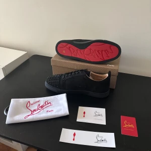 Christian Louboutin svarta sneakers med nitar - Säljer ett par svarta sneakers från Christian Louboutin med ikonisk röd sula och svarta nitar på tån. Skorna är i mocka och har snörning. Perfekt för dig som vill sticka ut med en lyxig och edgy stil. Originalförpackning och dustbag medföljer.