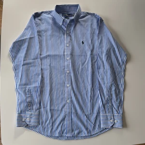 Ralph Lauren Blåvit randig skjorta  M - Klassisk blå och vit randig skjorta från Ralph Lauren i modell Classic Fit. Skjortan har långa ärmar, knappar framtill och på manschetterna samt den ikoniska broderade loggan på bröstet. Tillverkad i mjuk bomull och passar perfekt till en stilren look.