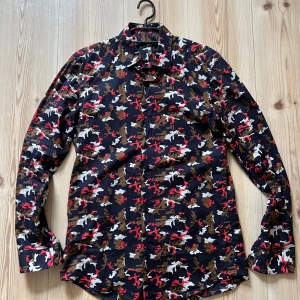 Camouflage skjorta från Hugo Boss - Snygg slim fit skjorta från Hugo Boss med färgstarkt camouflagemönster i marinblått, rött, vitt och brunt. Skjortan har långa ärmar, klassisk krage och knäppning framtill. Tillverkad i mjuk bomull med italienskt tyg. Perfekt för dig som vill sticka ut.