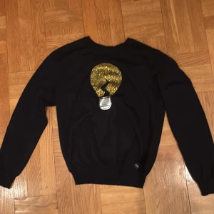 Svart Fendi tröja med glittrig detalj - Svart tröja från Fendi med lång ärm och rund hals. Framsidan har en stor glittrig guld- och silverapplikation i form av en glödlampa. Tröjan är i mjukt material och har en klassisk passform. Perfekt för dig som vill sticka ut med en unik designerpiece. Tröjan är unik och i riktigt bra skick, den är såklart äkta. Priset är inte hugget i sten som kom med förslag så kommer vi överens om något 