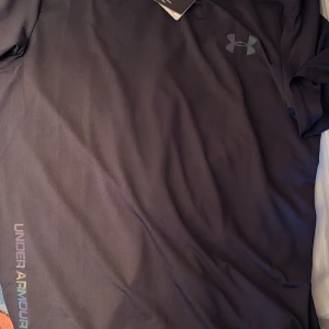 Svart Under Armour HeatGear t-shirt - Under Armour HeatGear t-shirt i svart med diskret logga på bröstet och färgglad text längs sidan. Tillverkad i lätt polyester som andas och håller dig sval. Passformen är slim och den har korta ärmar, perfekt för träning och aktiva dagar.