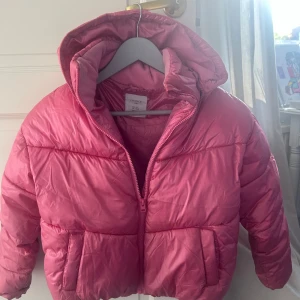 Rosa pufferjacka från Lindex, stl 146 - Rosa varm barn vinterjacka från Lindex med huva och dragkedja. Jackan har två stora fickor framtill och är fodrad med mjuk fleece på insidan av kragen. 