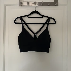 Svart sporttopp från Stronger - Svart sporttopp från Stronger med smala axelband som korsas i ryggen och justerbara detaljer. Toppen är croppad och har en stilren design med diskret logga framtill. Perfekt för träning och tillverkad i mjukt, stretchigt syntetmaterial. 💋