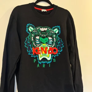 Svart Kenzo tröja med tigerbrodyr - Svart tröja från Kenzo med ikonisk tigerbrodyr i grönt, blått och vitt samt röd Kenzo-text på bröstet. Rund halsringning och långa armar.