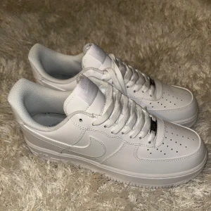 Nike Air Force 1 helvita sneakers - Klassiska Nike Air Force 1 sneakers i helvitt skinn med vit sula och snörning. Ikonisk design med perforerad tå, broderad Nike Air-logga på hälen och diskret silverdetalj vid snörningen. Perfekta för en clean och stilren look.