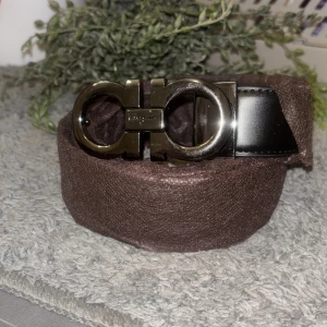 Svart skärp från Salvatore Ferragamo - Säljer ett svart skärp från Salvatore Ferragamo med ikoniskt dubbel-Gancini spänne i silverfärgad metall. Bältet är tillverkat i äkta skinn och har en stilren, klassisk design som lyfter din outfit. Perfekt för dig som gillar exklusiva accessoarer.