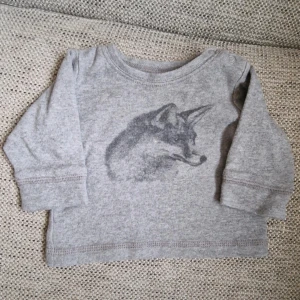 Grå långärmad tröja med rävtryck - Grå långärmad tröja i mjuk bomull med ett snyggt tryck av en räv på bröstet. Tröjan har rund halsringning och knapp på axeln för enkel påklädning. Lite ytludd har jag sett men inga andra skavanker.
