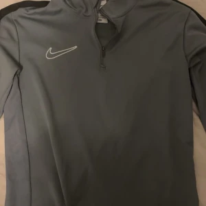 Grå Nike Dri-Fit half zip tröja XS - Nike Dri-Fit tröja i grått med halv dragkedja och lång ärm. Perfekt för träning eller chill, med klassisk Nike-logga på bröstet. Materialet är syntet och designen är clean och sportig.