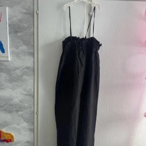 Svart långklänning med smala axelband - En svart långklänning med smala axelband och rynkad överdel. Klänningen har en rak och luftig passform, perfekt för varma dagar. Enkel och stilren design som är lätt att matcha med accessoarer. Ny med prislapp