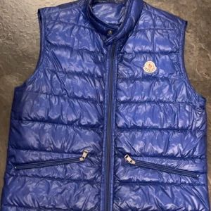 Blå dunväst från Moncler - Snygg blå dunväst från Moncler med glansig finish och klassisk logga på bröstet. Västen har hög krage, två fickor med dragkedja och quiltad design. Perfekt för lager-på-lager och streetstyle.