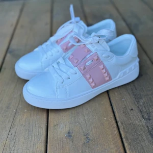 Valentino Rockstud sneakers vit/rosa - Snygga Valentino Rockstud sneakers i vitt skinn med rosa detaljer och ikoniska nitar. Skorna har rund tå, platt sula och breda vita snören. Perfekt för dig som vill sticka ut med en lyxig och trendig look. Märkeslogga på insidan och coola detaljer på sulan. Vet ej om dessa är äkta, köpte på Vinted men var för små. Gav 2000 kr men säljer för 1500 kr prutat och klart.
