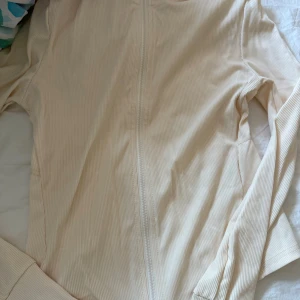 Beige ribbad hoodie med dragkedja - Snygg beige hoodie med ribbad struktur och hel dragkedja framtill. Tröjan har långa ärmar och en stilren look som passar perfekt till träning eller vardag. Enkel att matcha med jeans eller leggings för en avslappnad stil.