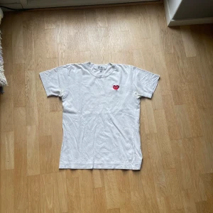 Vit Comme des Garçons Play t-shirt - Vit t-shirt från Comme des Garçons Play med det ikoniska röda hjärtat broderat på bröstet. Klassisk passform och rund hals. Tillverkad i mjuk bomull, perfekt för en clean och stilren look. Passar s/xs
