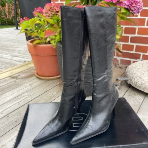 Svarta stövlar med klack från Bianco - Säljer ett par svarta höga stövlar från Bianco med spetsig tå och smal klack. Skorna har dragkedja på insidan för enkel påtagning och är köpta på 90-talet. Defekt uppkommer genom avflagnad av färg. 