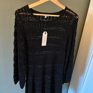 Svart stickad klänning från Escape - Svart stickad klänning från Escape med lång ärm och snyggt virkat mönster. Klänningen har rund halsringning och är perfekt för dig som gillar en avslappnad men ändå stilren look. Perfekt till stranden över en bikini.