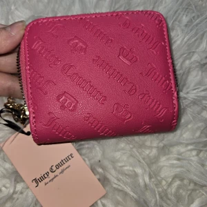 Rosa plånbok från Juicy Couture - Superfin rosa plånbok från Juicy Couture med präglad logga och små kronor över hela utsidan. Plånboken har guldfärgad dragkedja, ett litet hjärtformat metallhänge och insida med svartvitt logomönster. Perfekt storlek för kort och mynt, riktigt snygg accessoar!