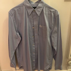 Ljusblå skjorta från Fred Perry - Snygg ljusblå skjorta från Fred Perry med klassisk krage och knäppning framtill. Skjortan har lång ärm, bröstficka med broderad logga och är tillverkad i mjuk bomull. Perfekt för en clean och stilren look.