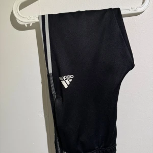 Svarta Adidas träningsbyxor med ränder - Svarta träningsbyxor från Adidas med klassiska vita ränder längs sidorna och Adidas-logga på låret. Byxorna har elastisk midja och är tillverkade i ett mjukt syntetmaterial som passar perfekt för sport och träning.