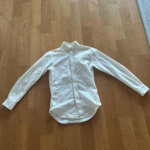 Vit skjorta från Ralph Lauren - Klassisk vit skjorta från Ralph Lauren med broderad logga på bröstet. Skjortan har lång ärm, knappar framtill och en snygg krage. Tillverkad i mjuk bomull och har en tidlös, clean look som passar till allt.