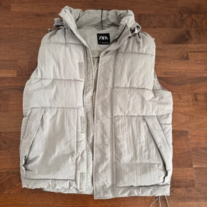 Ljusgrå dunväst från Zara - Ljusgrå dunväst från Zara med quiltad design och dragkedja framtill. Västen har hög krage med dragsko och två snedställda fickor. 