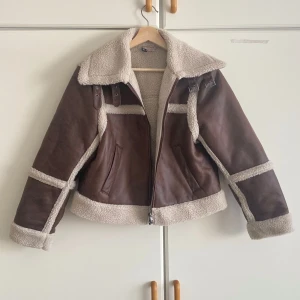 Brun teddyjacka med fuskpälsdetaljer - Säljer en brun teddyjacka med krämvita fuskpälsdetaljer och bred krage. Jackan har dragkedja framtill, två fickor och coola spännen vid halsen. Perfekt för dig som vill ha en trendig och varm look under kalla dagar.