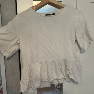 Vit volangblus från Bik Bok - Supersöt vit blus från Bik Bok i bomull med volang nertill. Modellen har rund halsringning och korta ärmar. Perfekt för dig som gillar en enkel fin topp.