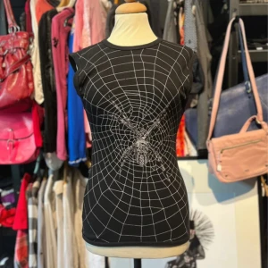 Svart topp med spindelnätsmönster  - Cool svart topp från Zip It London med ett stort vitt spindelnät och en detaljerad spindel tryckt både fram och bak. Ärmlös och figurnära modell i mjuk bomull, perfekt för dig som gillar edgy och unika prints.