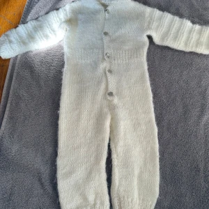 Vit stickad byxdress med huva - Mysig vit stickad byxdress med långa ärmar, huva och knappar framtill. Perfekt för kalla dagar och superbekväm att chilla i. Byxdressen är ribbstickad vid ärmslut och benslut för extra komfort.