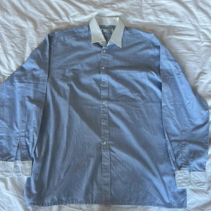 Blårandig skjorta Nordiska Kompaniet - Klassisk blårandig skjorta från Nordiska Kompaniet med vit krage och vita manschetter. Skjortan har knappar framtill, bröstficka och lång ärm. Snygg och stilren med diskreta tunna ränder och relaxed fit. 